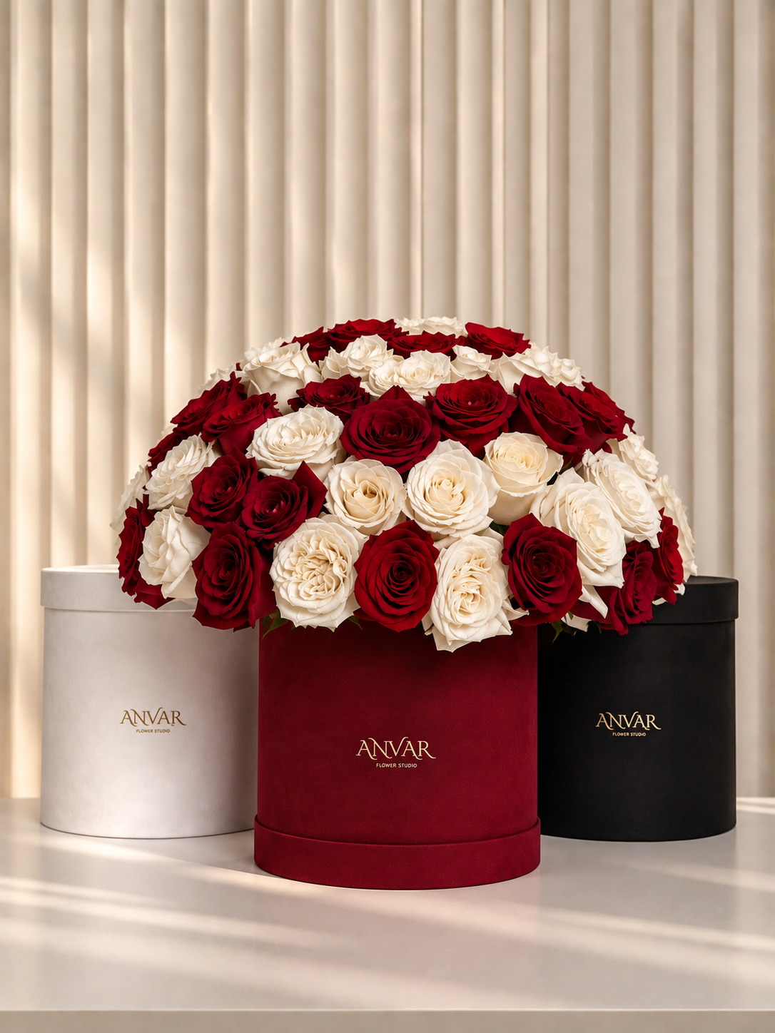 A large dome arrangement of red and white roses displayed in a red round flower box, with two cylindrical Anvar gift boxes on either side against a cream pleated backdrop.
